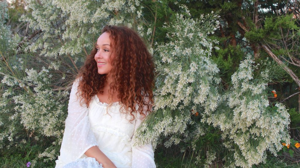 Author coach and podcast host Cynthia Occelli sitting on the ground in a white dress in front of trees and plants